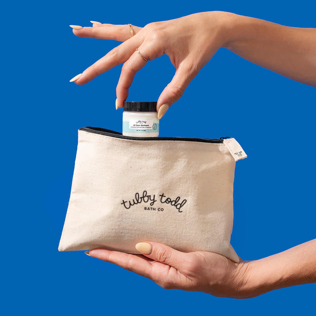 A hand holding the 1oz All Over Ointment jar from the the canvas travel bag.